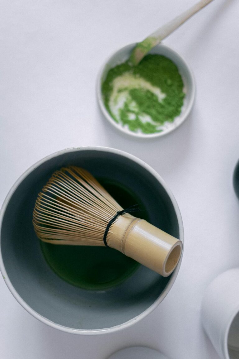 A flat lay of matcha tea preparation with a bamboo whisk and powder on a white background.